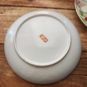 May include: A pale blue ceramic plate with a white rim, seen from below. A small, red and gold stamp is visible near the centre. The plate rests on a wooden surface.