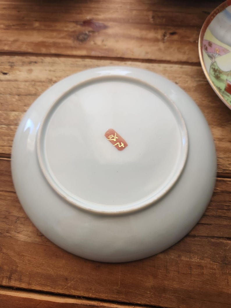 May include: A pale blue ceramic plate with a white rim, seen from below. A small, red and gold stamp is visible near the centre. The plate rests on a wooden surface.