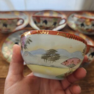 May include: A hand-painted vintage teacup with a landscape design featuring mountains, trees, and a pink fan. The cup has a gold rim and is part of a set. Other teacups and saucers are visible in the background.