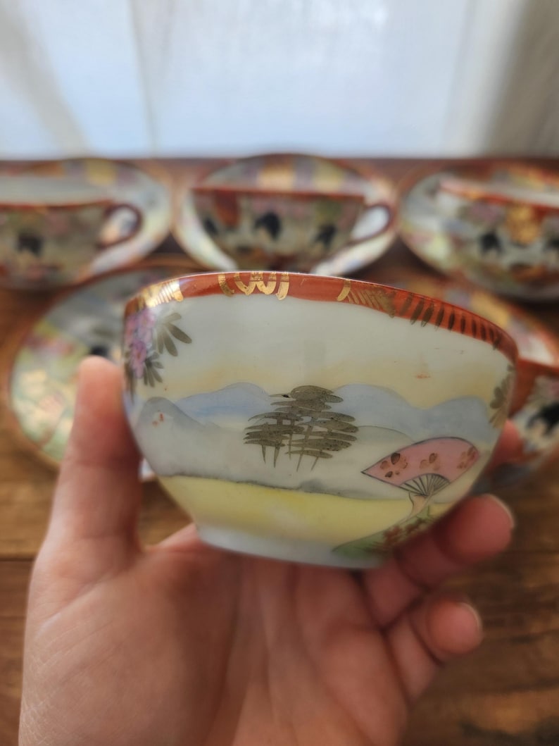 May include: A hand-painted vintage teacup with a landscape design featuring mountains, trees, and a pink fan. The cup has a gold rim and is part of a set. Other teacups and saucers are visible in the background.