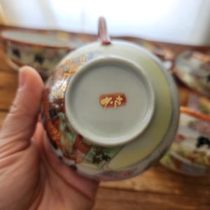 May include: A Japanese teacup with a handle, showcasing detailed floral and gold patterns. The cup's interior is pale green, and the base has a red stamp with Japanese characters. Other teacups are visible in the background, with a wooden surface.