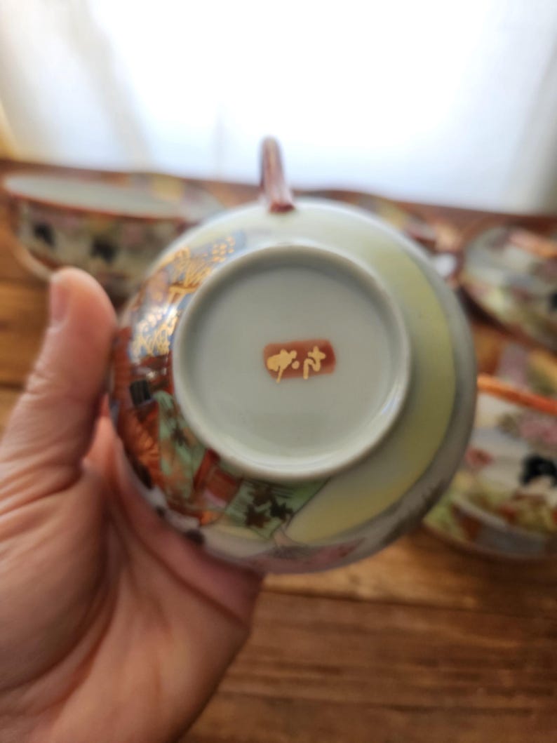 May include: A Japanese teacup with a handle, showcasing detailed floral and gold patterns. The cup's interior is pale green, and the base has a red stamp with Japanese characters. Other teacups are visible in the background, with a wooden surface.