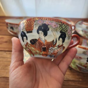 May include: A hand-painted porcelain teacup with a red handle and rim. The cup features a detailed illustration of three women in traditional Japanese attire, set against a backdrop of flowers and gold accents. The teacup is part of a set.