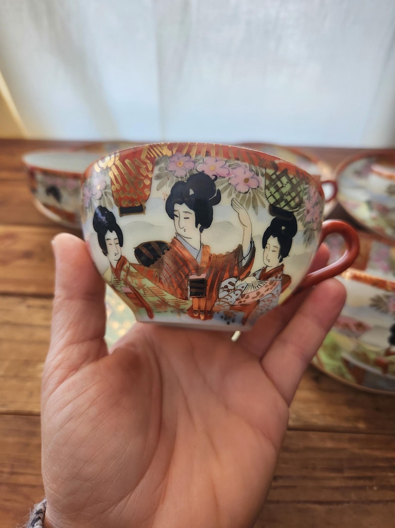 May include: A hand-painted porcelain teacup with a red handle and rim. The cup features a detailed illustration of three women in traditional Japanese attire, set against a backdrop of flowers and gold accents. The teacup is part of a set.