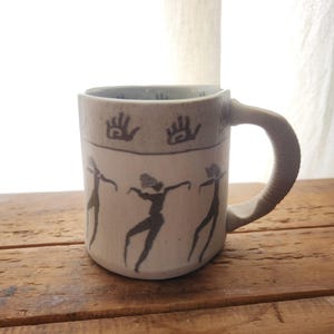 Puede incluir: Una taza de cerámica con exterior beige claro y asa curvada. La taza presenta siluetas negras de figuras danzantes y huellas de manos. La taza está sobre una superficie de madera.
