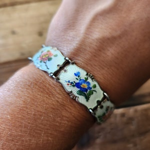 Puede incluir: Un brazalete de plata con una cadena delicada y detalles de flores de esmalte. Las flores están pintadas en tonos de azul, rosa y amarillo sobre un fondo blanco.