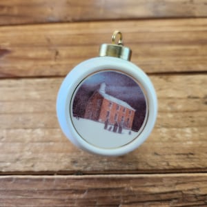 May include: White Christmas ornament with a gold-colored metal cap. The ornament features a circular image of a red brick building in a snowy landscape. The image is framed with a gold border. A festive holiday decoration.