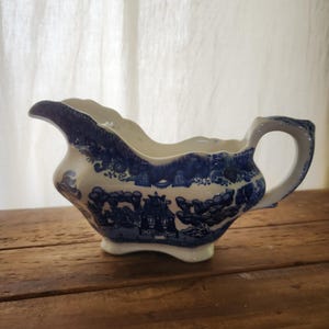 May include: A blue and white ceramic gravy boat with a handle and spout. The boat features a detailed landscape design in blue against a white background. The gravy boat is placed on a wooden surface.