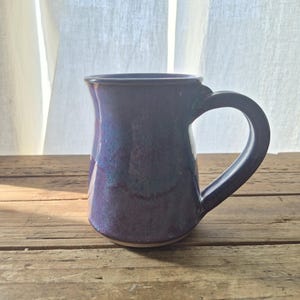May include: A ceramic mug with a glossy finish featuring a blend of deep purple, blue, and magenta tones. The mug has a wide, curved handle on the right side and a slightly flared rim. The base of the mug shows a thin, unglazed light-colored rim.