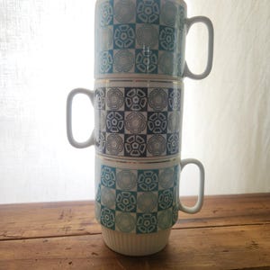 May include: Three stacked ceramic mugs with a geometric pattern in shades of blue and white. Each mug has a white handle and a gold rim. The mugs are arranged on a wooden surface.