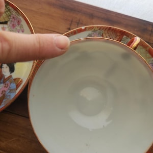 May include: Close-up of a white teacup with a gold rim and a decorative saucer. The saucer features a painted design of a woman in traditional Japanese attire, with floral and geometric patterns in red, pink, and gold.