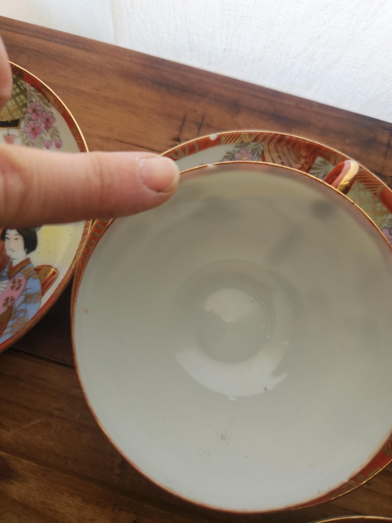 May include: Close-up of a white teacup with a gold rim and a decorative saucer. The saucer features a painted design of a woman in traditional Japanese attire, with floral and geometric patterns in red, pink, and gold.