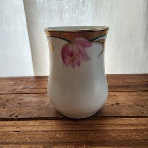 May include: A white ceramic vase with a gold rim and a pink tulip design. The vase has a rounded shape and is set on a wooden surface. The tulip is painted in shades of pink and green.