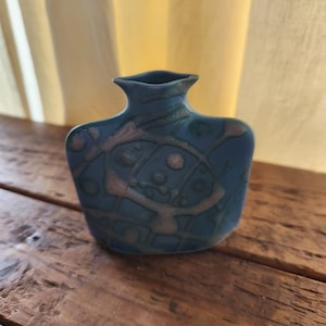 May include: A small, blue ceramic vase with a unique, abstract design in pink and green. The vase has a square base and a flared neck. The design features geometric shapes and lines. The vase is sitting on a wooden surface.