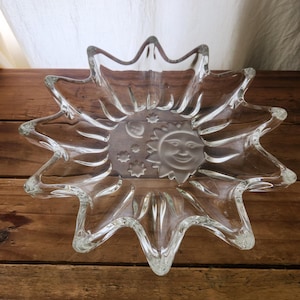May include: A clear glass bowl with a starburst design. The bowl features a smiling sun and crescent moon with stars in the center. The bowl is made of clear glass and sits on a wooden surface.