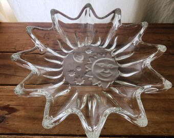 May include: A clear glass bowl with a starburst design. The bowl features a smiling sun and crescent moon with stars in the center. The bowl is made of clear glass and sits on a wooden surface.