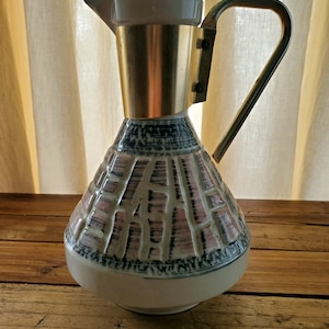 May include: A white ceramic pitcher with a gold rim and handle. The pitcher has a geometric pattern in shades of pink, black, and white.