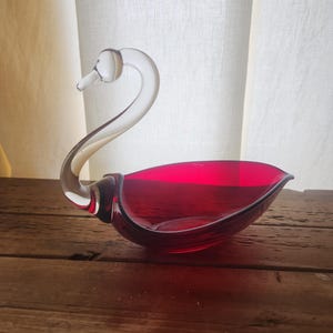 Puede incluir: Un cuenco en forma de cisne de cristal rojo rubí con cuello y cabeza de cristal transparente. El cuenco está sobre una superficie de madera, con una cortina blanca al fondo. El cuello del cisne se curva con gracia y el cuenco tiene una forma suave y redondeada.