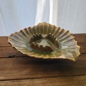 May include: A vintage ashtray with a unique leaf-like design. The ceramic ashtray features a central area for holding cigarettes and a wide, ruffled edge. The color is a mix of green and brown with gold accents, giving it a retro aesthetic.