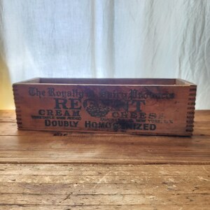May include: A vintage wooden box with the text "The Royally Dairy Products REGT Cream Cheese" printed on the front. The box is brown and sits on a wooden surface. The text also includes "Doubly Homogenized".