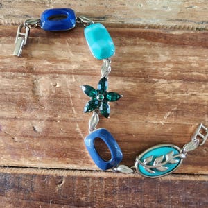 May include: A silver-toned bracelet with blue, turquoise, and green accents. The bracelet features a dark blue square, a turquoise rectangle, a green flower, another dark blue square, and an oval with a leaf design.