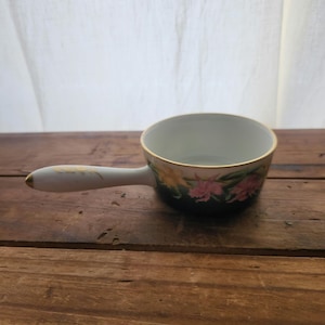 May include: A small white porcelain sauce pot with a floral design. The pot has a gold rim and a handle. The floral design features pink, yellow, and green flowers on a dark green background.