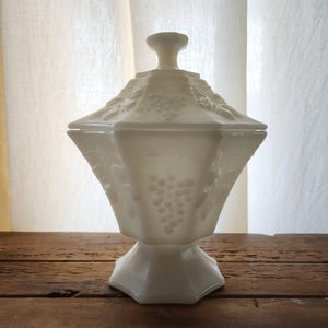 May include: A white, lidded candy dish with a pedestal base. The dish features a geometric shape with embossed grape and leaf designs. The lid has a small, rounded handle. The dish is set on a rustic wooden surface.