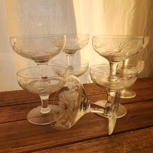 May include: A set of clear, etched glass champagne coupe glasses. Each glass has a floral design and a short stem. The glasses are arranged on a wooden surface, with one glass lying on its side.