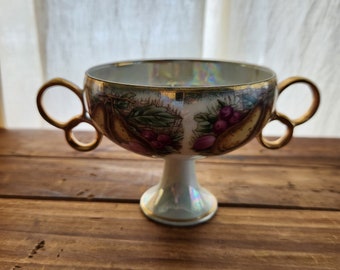 May include: A white porcelain cup with a gold rim and a floral design. The cup has two handles and sits on a pedestal.