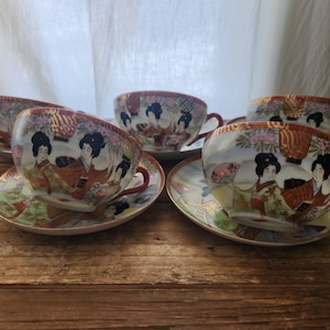 May include: Set of five vintage Japanese porcelain teacups and saucers. Each cup and saucer features a hand-painted design of geishas in traditional attire, with floral and landscape elements. The cups have a red rim and handle.