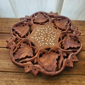Puede incluir: Una bandeja redonda de madera tallada intrincadamente con un diseño floral y de hojas. La bandeja está hecha de madera marrón oscuro y presenta un patrón central con incrustaciones y acentos de color claro. Los bordes están adornados con hojas talladas y motivos florales.