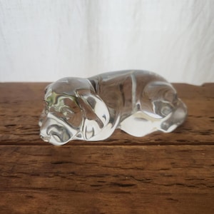 Puede incluir: Una figura de perro de cristal transparente en posición de descanso. La escultura tiene detalles precisos, incluyendo las orejas, ojos y nariz del perro. El cristal es transparente y está sobre una superficie de madera.