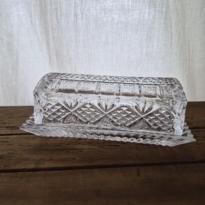 May include: A rectangular clear glass butter dish featuring a detailed pressed diamond and crosshatch pattern on the lid and base. The lid has a flat top with a geometric border, while the base has a serrated edge design around the perimeter.