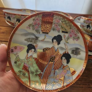 May include: A small, round, decorative plate with a traditional Japanese design. The plate has a red border and depicts three women in colourful kimonos, holding fans, with a backdrop of flowers and mountains. Matching bowls are visible.