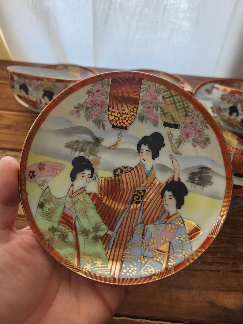 May include: A small, round, decorative plate with a traditional Japanese design. The plate has a red border and depicts three women in colourful kimonos, holding fans, with a backdrop of flowers and mountains. Matching bowls are visible.