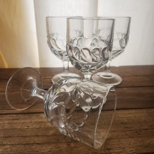 May include: Four clear, stemmed wine glasses with a decorative pattern. One glass is lying on its side on a wooden surface. The other three glasses are upright in the background. The glasses are empty.