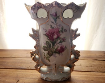 May include: A white porcelain vase with gold accents and a floral design featuring pink roses. The vase has a unique, ornate design with cutouts and a scalloped rim.