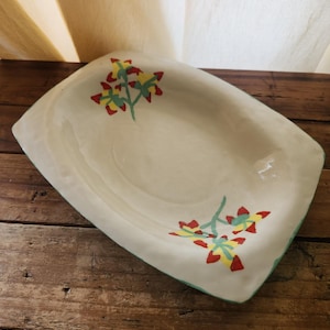 May include: A white ceramic rectangular serving dish with a green rim and a floral design featuring red, yellow, and green flowers.