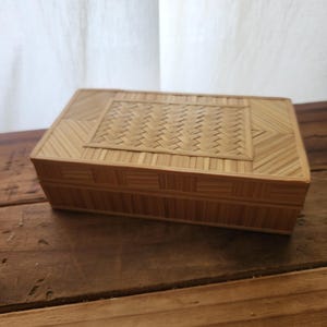 May include: A rectangular bamboo box with a woven top and a natural finish. The box is made of thin strips of bamboo, with a woven pattern on the lid and a geometric design on the sides. The box is sitting on a wooden surface.