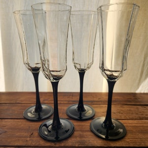 May include: Four clear glass champagne flutes with black stems. The flutes are arranged in a row on a wooden surface.