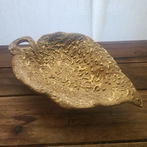 Puede incluir: Un plato de cerámica con forma de hoja con una superficie texturizada y un asa de bucle. El plato es de color marrón cálido y terroso, con reflejos más claros. El plato está sobre una superficie de madera.
