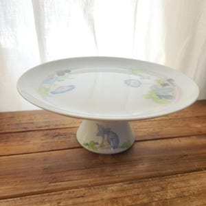 May include: White ceramic cake stand with a floral design in blue, green, and pink. The stand has a wide, flat top and a short, pedestal base. The cake stand is on a wooden surface.