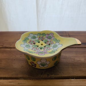 May include: Yellow ceramic tea strainer with a floral design. The strainer has a handle with a gold accent and is decorated with pink, white, and green flowers. The tea strainer is on a wooden surface.