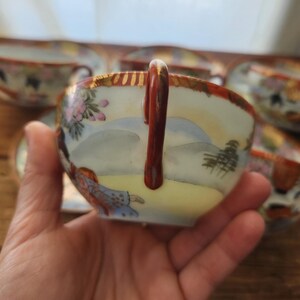 May include: A hand holding a porcelain teacup with a landscape design. The cup has a brown handle and a gold rim. The design includes pink flowers, a figure, and a mountain scene. Other cups are visible in the background.