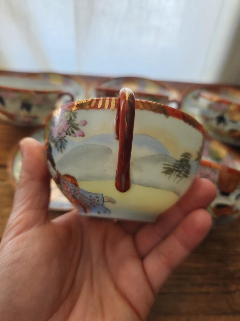 May include: A hand holding a porcelain teacup with a landscape design. The cup has a brown handle and a gold rim. The design includes pink flowers, a figure, and a mountain scene. Other cups are visible in the background.