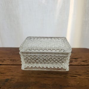 May include: A clear, rectangular glass trinket box with a diamond pattern. The box has a matching lid and sits on a wooden surface. The box is likely used for storing small items like jewelry or keepsakes.