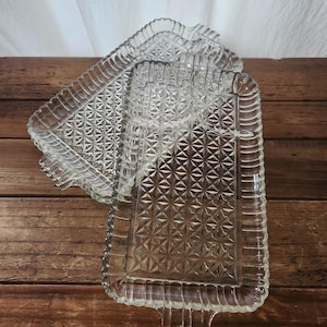 May include: Two clear glass serving trays with a diamond and starburst pattern. The trays have a rectangular shape with scalloped edges and small handles. They are stacked on a wooden surface.