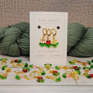 May include: A display of handmade tulip-shaped stitch markers with gold-colored clasps. The markers are red, pink, and yellow with green leaves. Two skeins of green yarn and a Pure Street card are in the background.