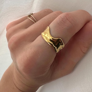 May include: A gold-toned ring with a wavy, abstract design. The ring is worn on a finger.