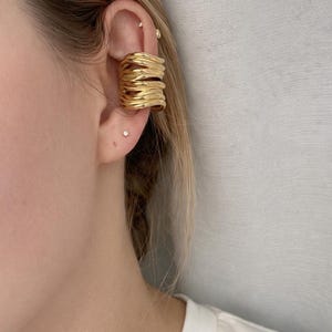 Puede incluir: Puño de oreja dorado con un diseño apilado y ondulado. El puño se envuelve alrededor de la oreja, creando un aspecto audaz y moderno. La imagen muestra el puño de oreja en la oreja de una persona, destacando su textura y brillo. Una joya minimalista.